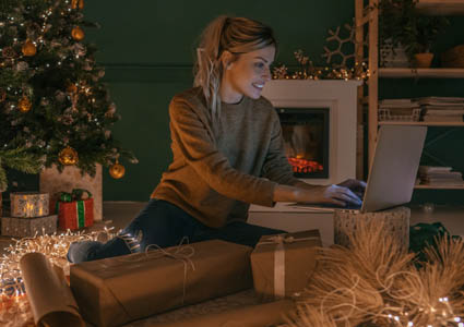 A woman on a laptop sitting in front of a decorated Christmas tree and recently wrapped gifts.